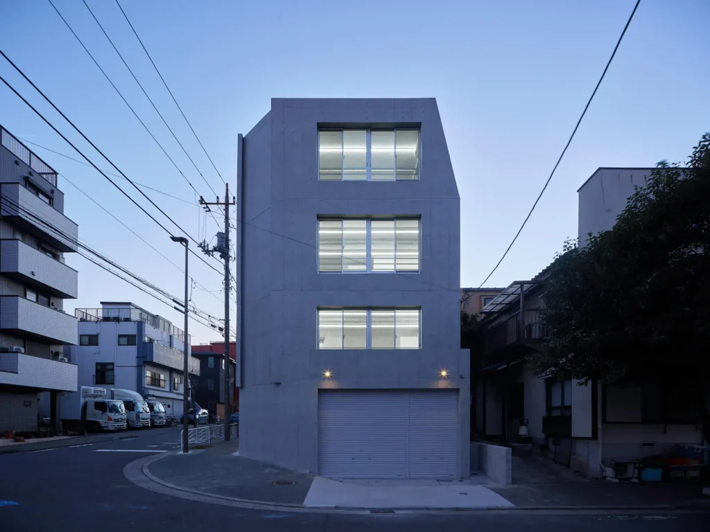 大開口窓から室内の光が溢れるRC造オフィスビルの東側正面外観（夕景）。コストダウンとデザイン性を両立。