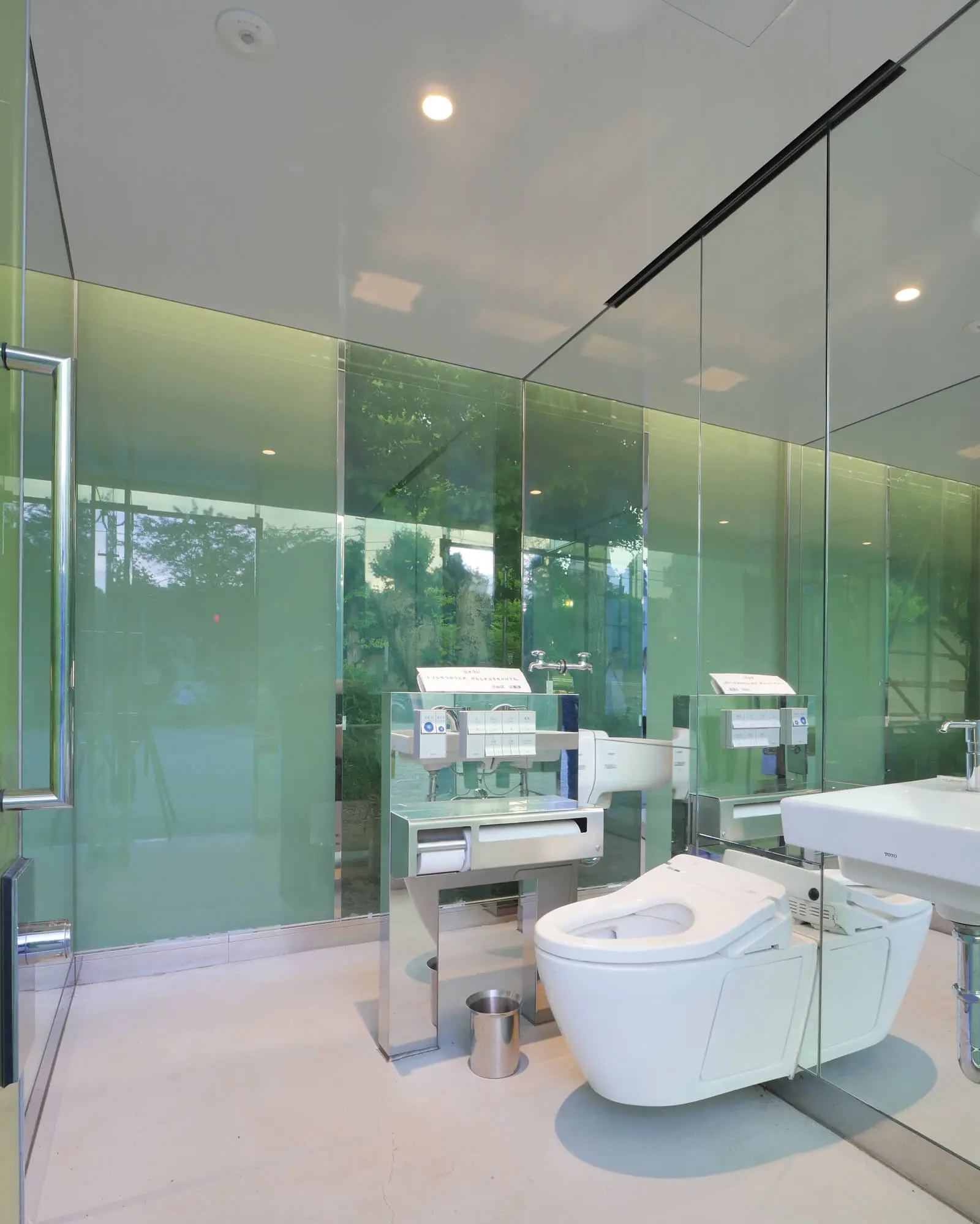 坂茂が手掛けた透明トイレの多目的トイレ（女子トイレ）内部。壁面のエポキシ樹脂とガラスが周囲の木々を映し出す、プライバシーと防犯を両立した空間。